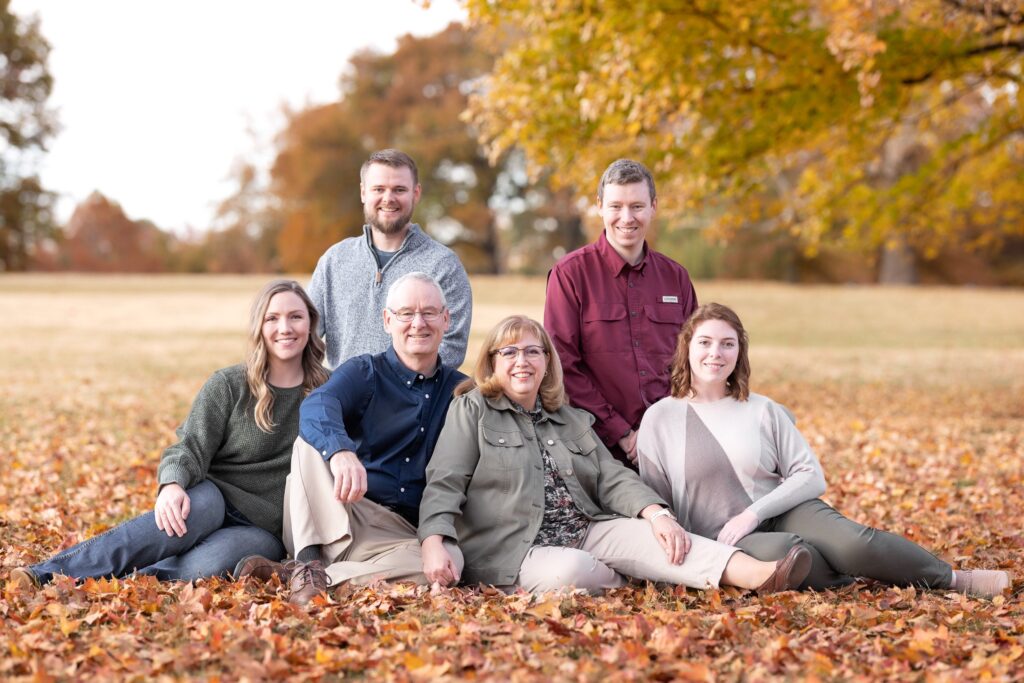 Family portrait in autumn setting, featuring six members seated on colorful fallen leaves, smiling and engaging with each other, reflecting familial support and connection.