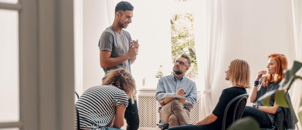 Group therapy session with a diverse group of young adults engaging in discussion, facilitated by a therapist, emphasizing support and emotional growth in a recovery setting.