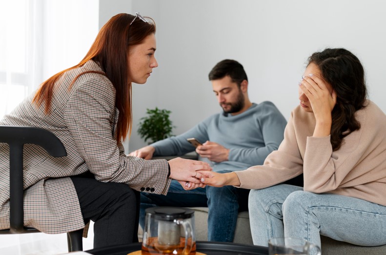 Supportive interaction between two women in a therapy session, one holding the other's hand, while a man sits in the background looking at his phone, emphasizing the importance of emotional support in mental health recovery.