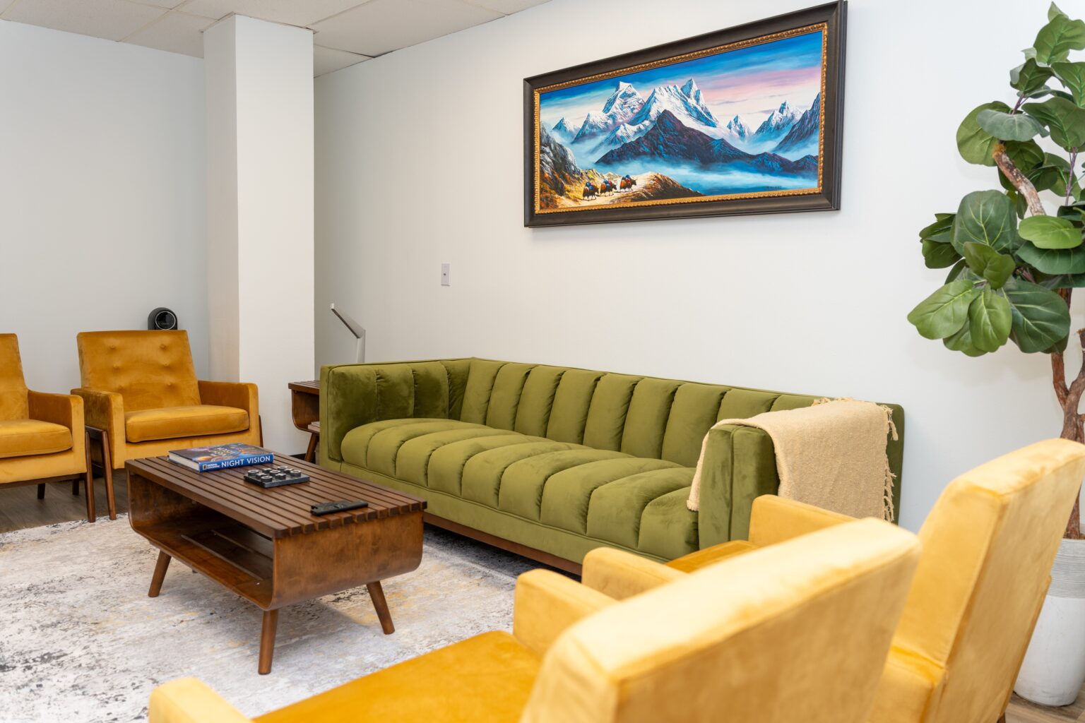 Cozy lobby area at Altura Recovery featuring a green velvet sofa, yellow armchairs, wooden coffee table with books, and a mountain landscape painting, designed for a welcoming atmosphere in addiction recovery support.