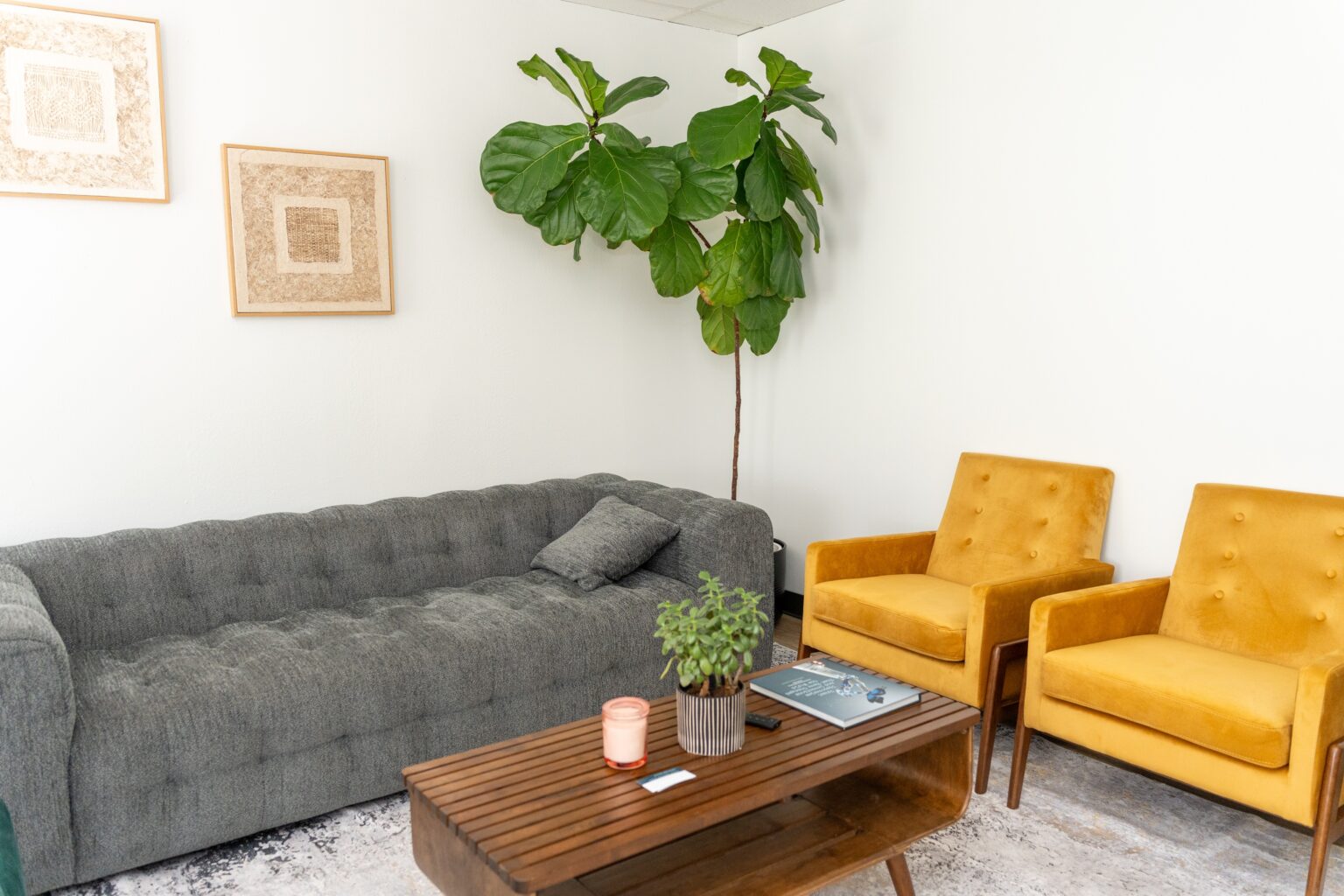 Living space at Altura Recovery featuring a gray tufted sofa, two yellow chairs, a wooden coffee table with a plant and a candle, and framed artwork on the wall, creating a welcoming environment for adult and teen recovery programs.
