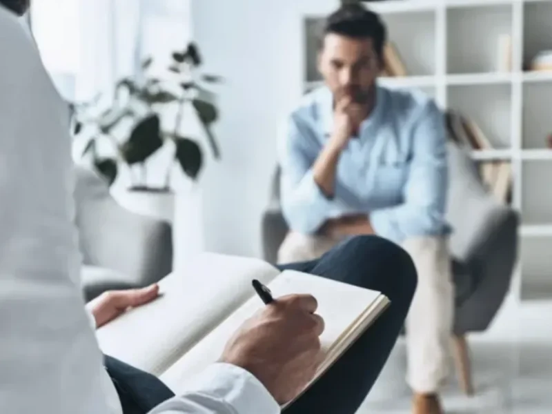 Person writing in notebook during therapy session with client, emphasizing mental health support and emotional recovery.