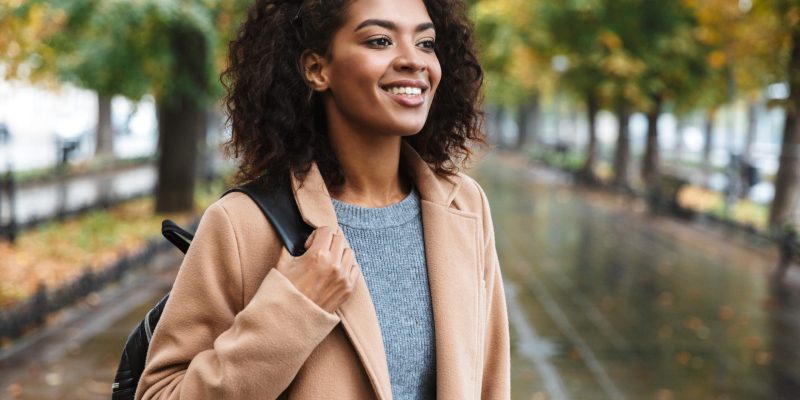 Beautiful young african woman wearing coat walking outdoors at the park, carrying backpack