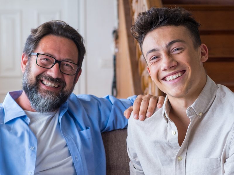 Happy father and son sitting on sofa at home. Cheerful father with teenage son sitting on couch in the living room of apartment. Portrait of father and son spending leisure time at home